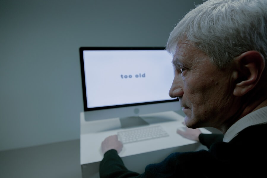 Un anciano sentado al lado de la computadora con un cartel que dice que es demasiado viejo para el trabajo al que se postuló